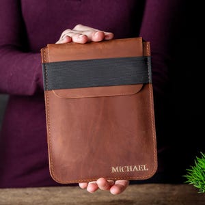 May include: A brown leather tablet sleeve with a black elastic band closure. The sleeve has a flap closure and the name "MICHAEL" is embossed in gold lettering. The sleeve is held by a person.