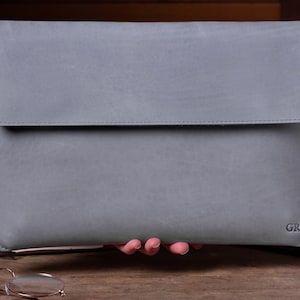 May include: A gray leather laptop sleeve with a flap closure and the initials "GR" embossed on the bottom right corner.