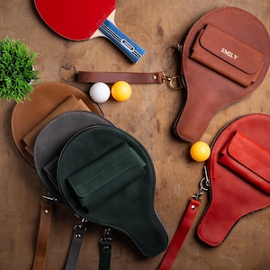 May include: A collection of leather ping pong paddle-shaped bags in various colors, including brown, red, and green. Each bag has a small wallet attached. A red and blue ping pong paddle, ping pong balls, and a small plant are also visible.