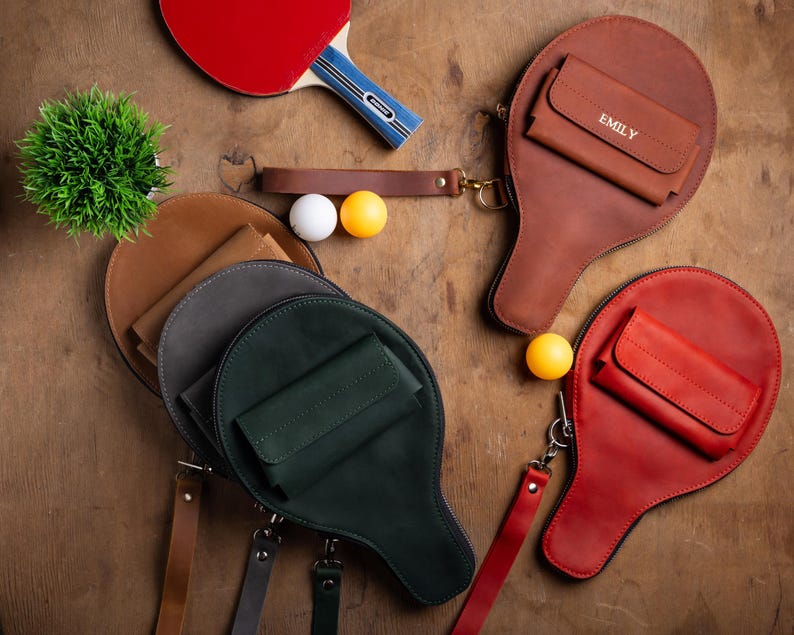 May include: A collection of leather ping pong paddle-shaped bags in various colors, including brown, red, and green. Each bag has a small wallet attached. A red and blue ping pong paddle, ping pong balls, and a small plant are also visible.