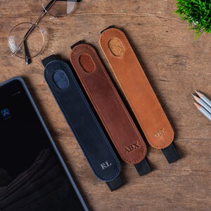 May include: Three leather pencil holders with personalized initials. The holders are brown, dark brown, and navy blue. The initials are in gold lettering. The holders are on a wooden surface.