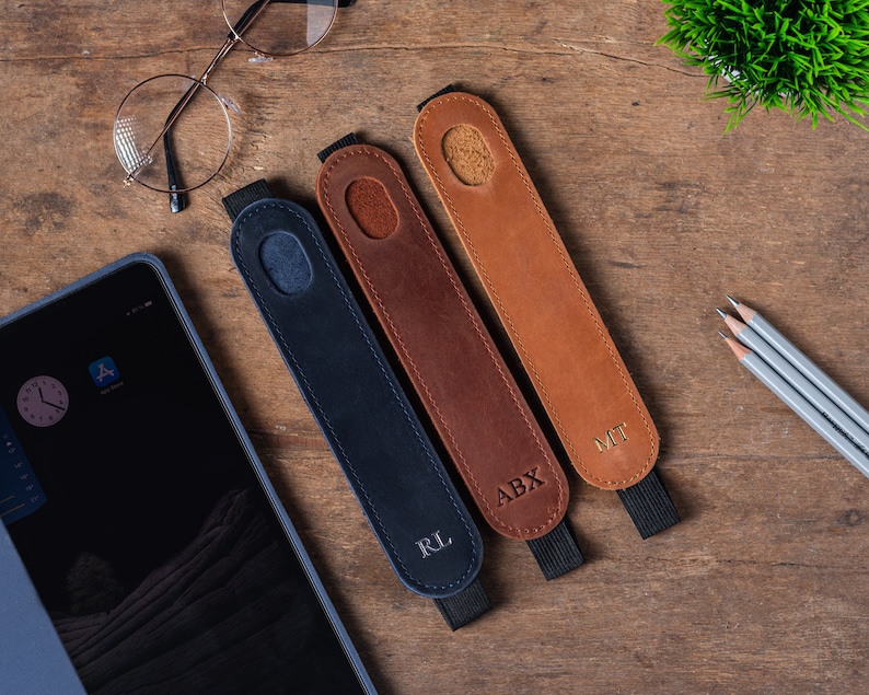 May include: Three leather pencil holders with personalized initials. The holders are brown, dark brown, and navy blue. The initials are in gold lettering. The holders are on a wooden surface.