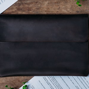 May include: A dark brown leather laptop sleeve with a flap closure. The sleeve is made of a high-quality leather and has a smooth, polished finish.