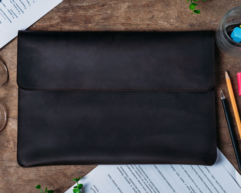 May include: A dark brown leather laptop sleeve with a flap closure. The sleeve is made of a high-quality leather and has a smooth, polished finish.