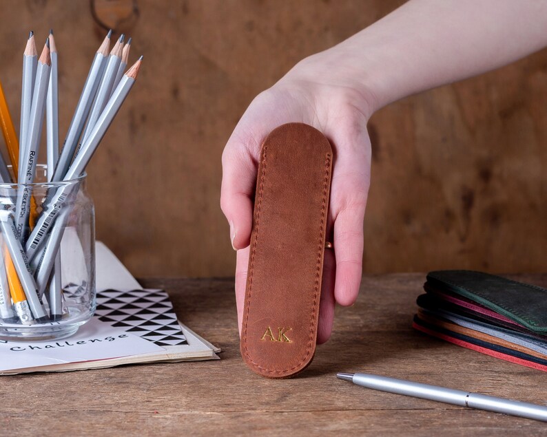 May include: A brown leather pencil case with the initials "AK" embossed in gold. The case is held in a hand.