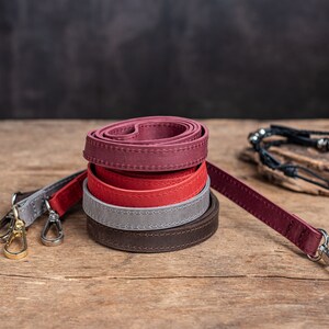 May include: Four leather straps in different colours, stacked on top of each other. The straps are brown, grey, red, and burgundy. The straps are rolled up and have stitching along the edges. The straps are on a wooden surface.