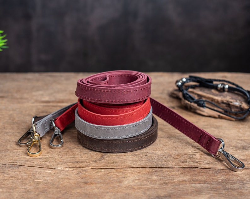 May include: Four leather straps in different colours, stacked on top of each other. The straps are brown, grey, red, and burgundy. The straps are rolled up and have stitching along the edges. The straps are on a wooden surface.