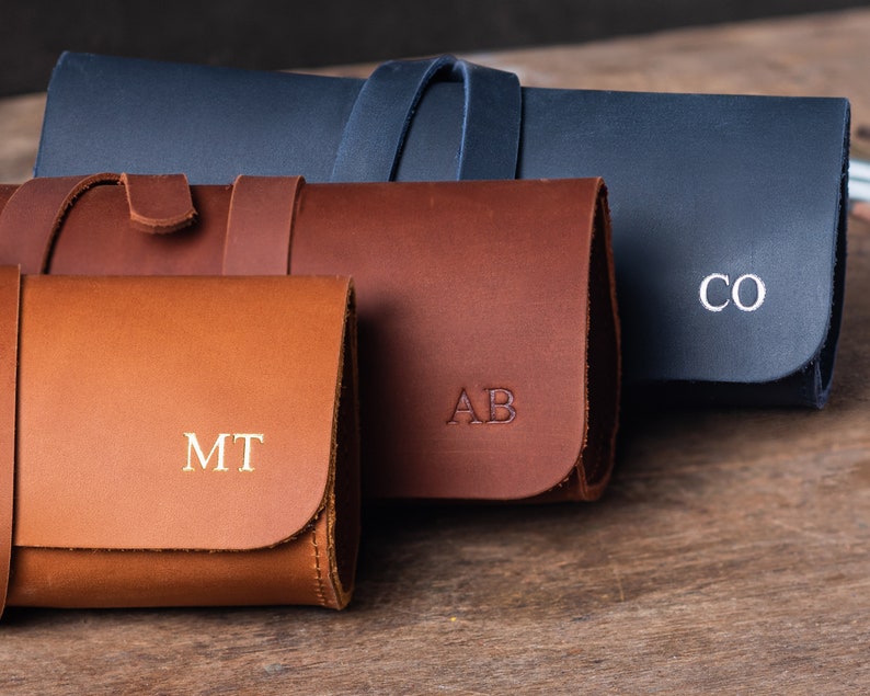May include: Three leather pouches with personalized initials. The pouches are brown, tan, and dark blue. The initials are "MT", "AB", and "CO".