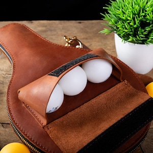 May include: A brown leather ping pong paddle case with a zipper, holding three white ping pong balls. The case has a pouch with a black velcro closure. A yellow ping pong ball is visible.