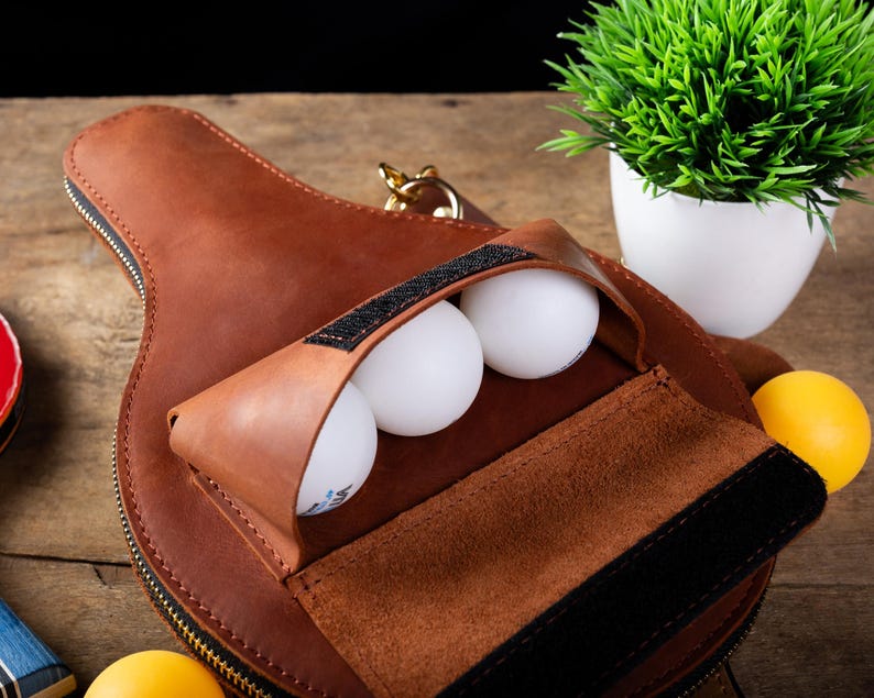 May include: A brown leather ping pong paddle case with a zipper, holding three white ping pong balls. The case has a pouch with a black velcro closure. A yellow ping pong ball is visible.