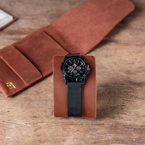 May include: A black wristwatch with a black nylon strap and a brown leather watch stand. The watch face has a black background with white numbers and hands, and a red second hand.