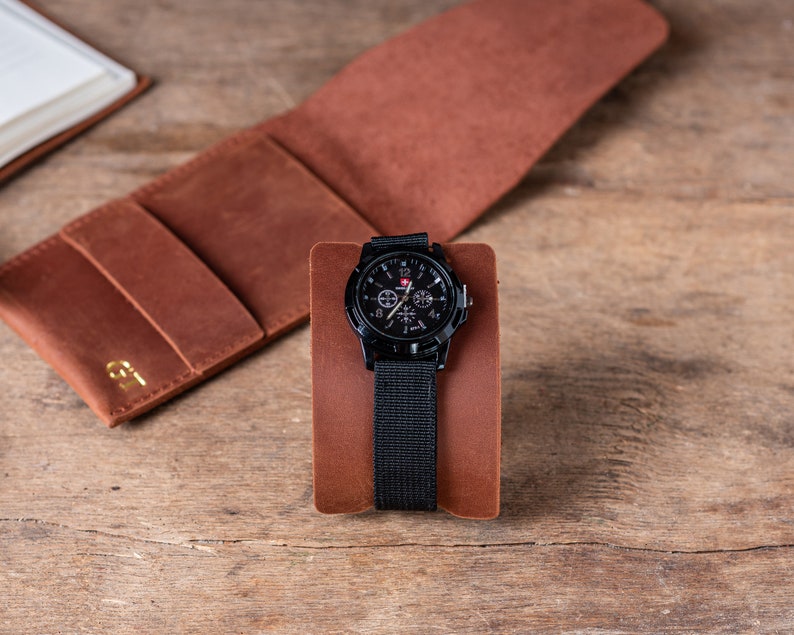 May include: A black wristwatch with a black nylon strap and a brown leather watch stand. The watch face has a black background with white numbers and hands, and a red second hand.
