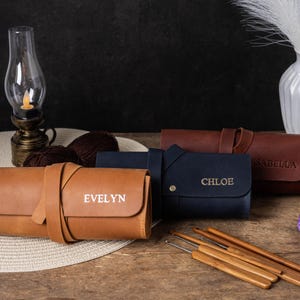 May include: Four brown leather needle cases with personalised names: Evelyn, Chloe, Isabella. The cases are rolled up and have a strap closure. There are also wooden knitting needles on a wooden table.