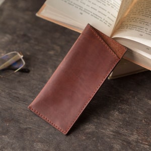 May include: A brown leather glasses case with a stitched edge. The case is lying on a wooden surface.