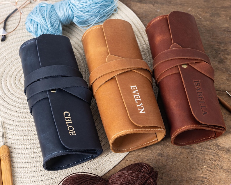 May include: Three leather needlepoint or knitting needle cases in different colours. The cases are rolled up and tied with leather straps. The cases are personalised with the names "Chloe", "Evelyn", and "Isabella".