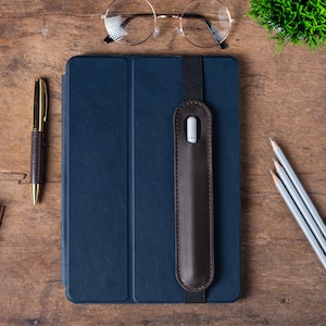 May include: A dark blue tablet case with a brown leather pencil holder attached. The case is on a wooden surface with a notebook, a pen, and three pencils.