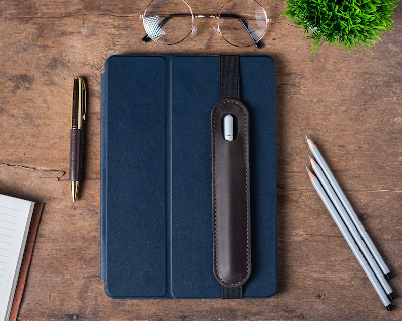 May include: A dark blue tablet case with a brown leather pencil holder attached. The case is on a wooden surface with a notebook, a pen, and three pencils.