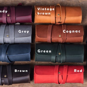 May include: Ten leather pencil cases in different colours, including burgundy, vintage brown, grey, cognac, blue, green, black, brown, red. Each case has a leather strap and a brass button.