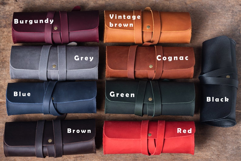 May include: Ten leather pencil cases in different colours, including burgundy, vintage brown, grey, cognac, blue, green, black, brown, red. Each case has a leather strap and a brass button.