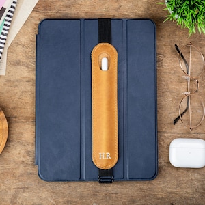 May include: A navy blue tablet case with a tan leather stylus holder, secured by a black elastic band. The stylus holder is embossed with the initials "HR". Gold-rimmed glasses and a white wireless charging case are also visible.