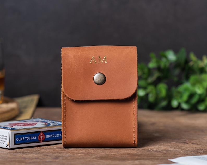 May include: A brown leather card case with a flap closure and a snap button. The case has the initials "AM" embossed in gold on the front.