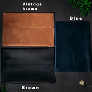 May include: Three leather laptop sleeves in different colors: vintage brown, blue, and brown. The sleeves are laid out on a black surface.