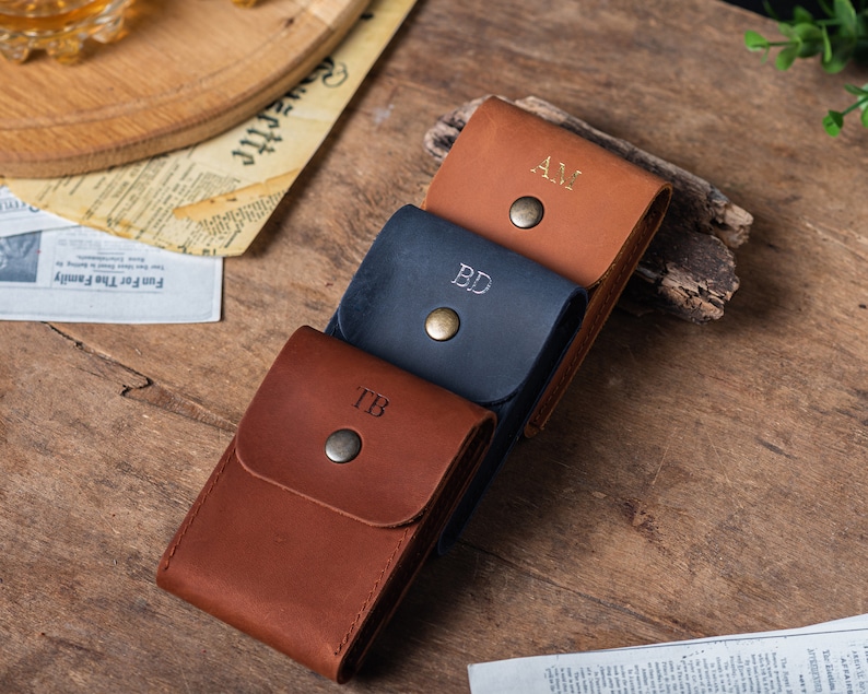 May include: Three leather card holders with initials 'TB', 'BD', and 'AJ' embossed on the front. The card holders are stacked on top of each other on a wooden surface.