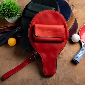 May include: A collection of leather ping pong paddle-shaped bags in red, green, blue, and brown. Each bag has a small pocket and a wrist strap. A ping pong paddle and balls are also visible.