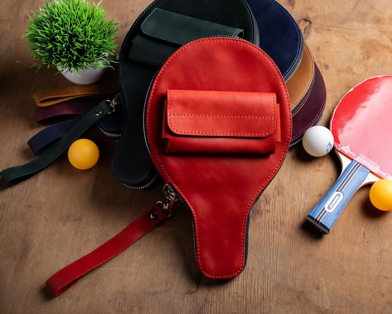 May include: A collection of leather ping pong paddle-shaped bags in red, green, blue, and brown. Each bag has a small pocket and a wrist strap. A ping pong paddle and balls are also visible.