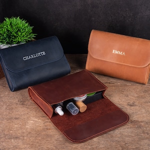 May include: Three leather pouches with personalised names, "Charlotte", "Emma", and one with the pouch open showing the interior with compartments and small bottles.