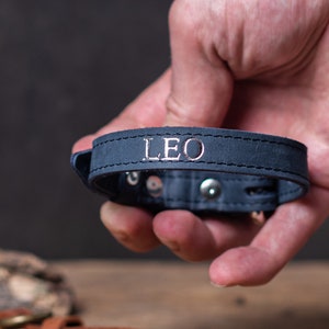 May include: A dark blue leather bracelet with silver stitching and the name "LEO" embossed in silver lettering.