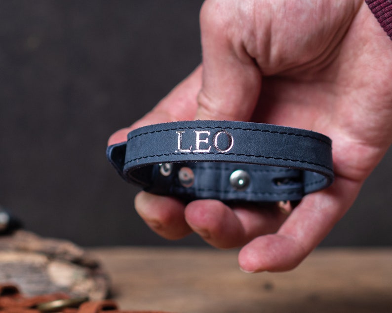 May include: A dark blue leather bracelet with silver stitching and the name "LEO" embossed in silver lettering.