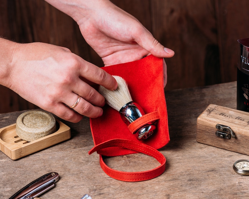 May include: A red leather shaving brush holder with a white shaving brush inside. The holder has a strap and is being held by a hand.