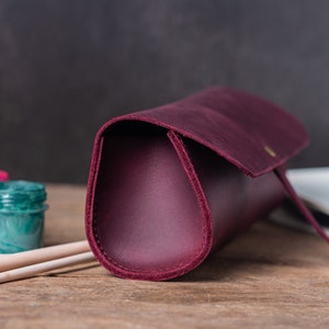 May include: A burgundy leather pencil case with a flap closure and a gold button. The case is lying on a wooden surface with two paintbrushes and paint jars in the background.