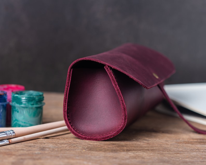 May include: A burgundy leather pencil case with a flap closure and a gold button. The case is lying on a wooden surface with two paintbrushes and paint jars in the background.