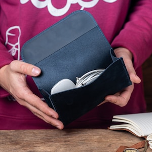 May include: A navy blue leather tech organizer, open to reveal a white mouse and white charging cables. The organizer is held in front of a person wearing a maroon sweatshirt. The background includes a wooden table and a notebook.