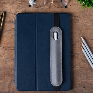 May include: A dark blue leather folio case with a gray elastic band and a gray felt pocket for a stylus. The case is closed and lying on a wooden surface.
