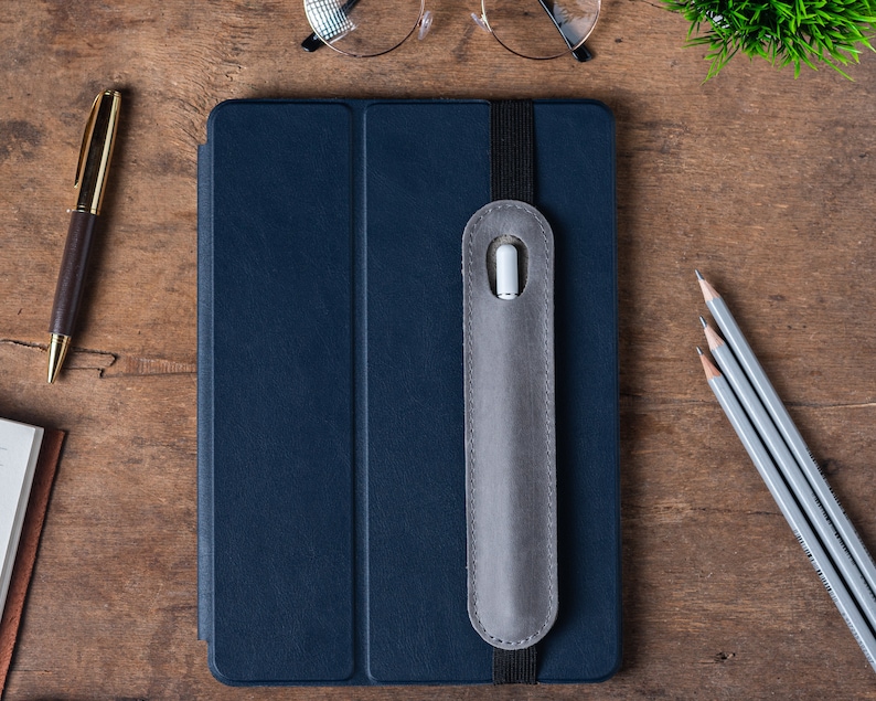 May include: A dark blue leather folio case with a gray elastic band and a gray felt pocket for a stylus. The case is closed and lying on a wooden surface.