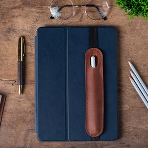 May include: A dark blue leather notebook cover with a brown leather pen holder attached. The cover has a flap that closes with a brown elastic band. The notebook is on a wooden surface with a gold pen and three pencils.