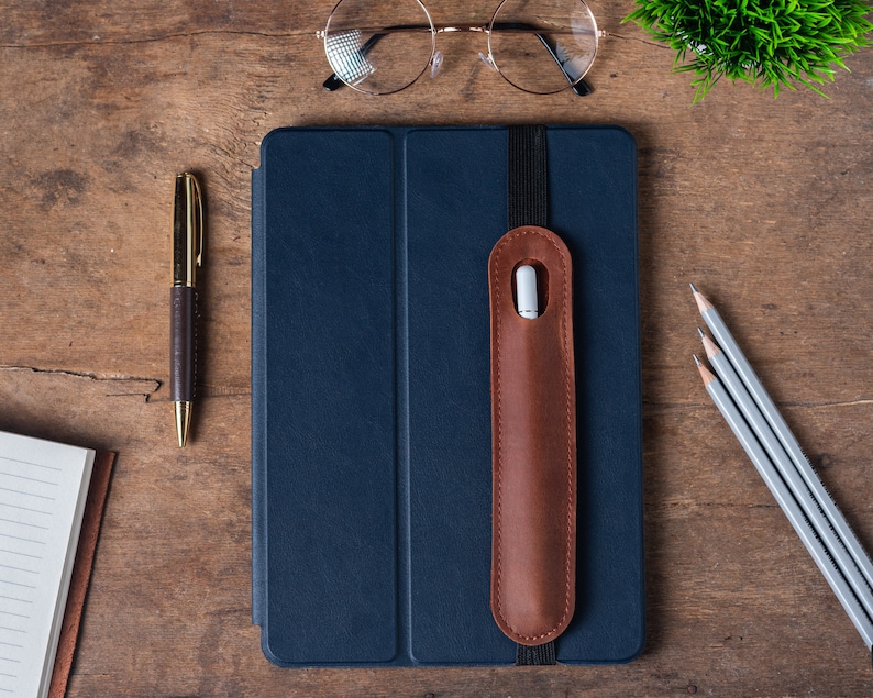 May include: A dark blue leather notebook cover with a brown leather pen holder attached. The cover has a flap that closes with a brown elastic band. The notebook is on a wooden surface with a gold pen and three pencils.