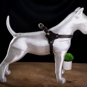 May include: A white Bull Terrier statue with a dark brown leather harness. The dog statue is standing on a wooden surface with a small green plant in a white pot in the background. The dog's tail is curved upwards.
