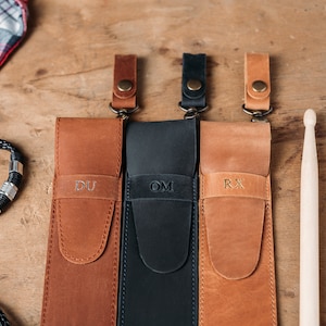 Puede incluir: Tres estuches de cuero para baquetas con iniciales personalizadas "DU", "OM" y "RX". Los estuches son de color marr&oacute;n, negro y marr&oacute;n, respectivamente. Los estuches tienen una correa y un lazo de metal. Dos baquetas de madera est&aacute;n a la derecha de la imagen.