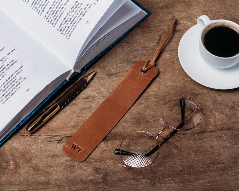May include: A brown leather bookmark with the initials "WT" embossed on it. The bookmark is lying on a wooden table next to a book, a pen, a cup of coffee, and a pair of round glasses.