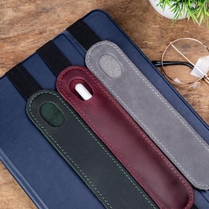 May include: Three leather stylus holders in dark green, burgundy, and gray, each with a contrasting stitch, rest on a blue tablet case. A white stylus is visible in the burgundy holder. The holders are attached to the case with black elastic bands.