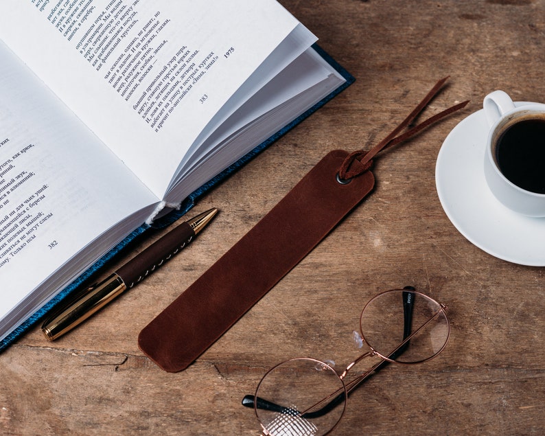 May include: A brown leather bookmark with a frayed edge, a gold pen, a pair of round glasses, a white coffee cup with black coffee, and a white saucer on a wooden surface.
