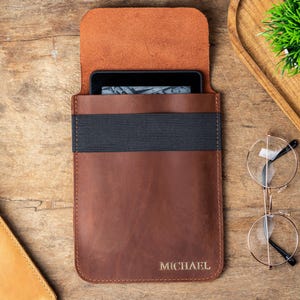 May include: A brown leather tablet sleeve with a black elastic band and the name "MICHAEL" embossed at the bottom. The sleeve holds a black tablet. The background includes a wooden surface, a small plant, and eyeglasses.