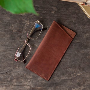 May include: A brown leather glasses case with a flap closure. The case is lying on a wooden surface next to a pair of silver-rimmed glasses.