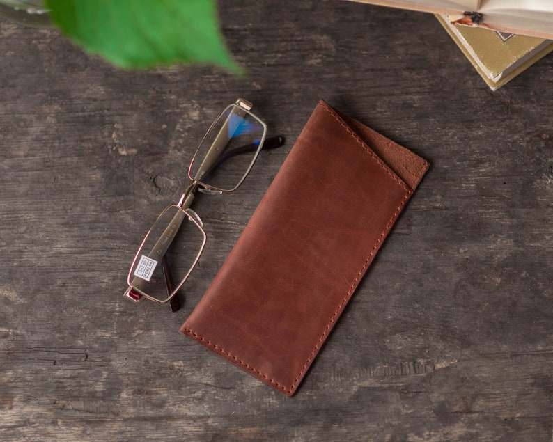 May include: A brown leather glasses case with a flap closure. The case is lying on a wooden surface next to a pair of silver-rimmed glasses.
