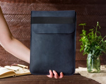Könnte beinhalten: Eine dunkelblaue Laptoptasche aus Leder mit einem schwarzen Gummibandverschluss. Die Tasche wird in einer Hand vor einem Holz Hintergrund gehalten.