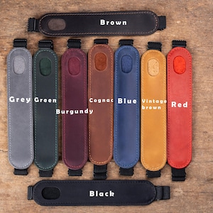 May include: A collection of leather camera strap pads in various colors, including grey, green, burgundy, cognac, blue, vintage brown, and red. The pads have black adjustable straps and are labeled with their respective colors. A brown and black strap pad is at the top, and a black strap pad is at the bottom.